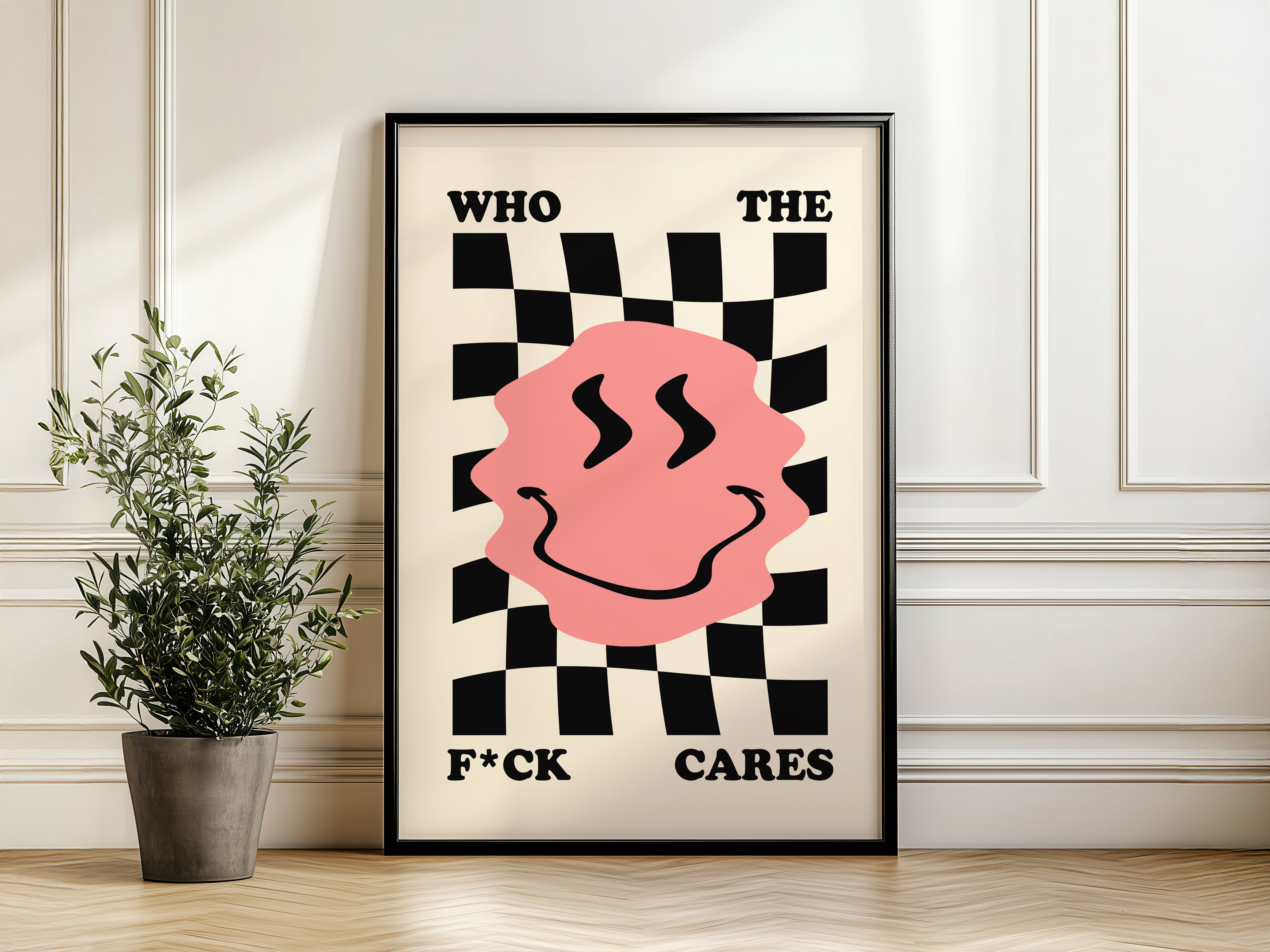 Framed poster with a pink face and text on a checkered background, placed on a wooden floor with a plant.